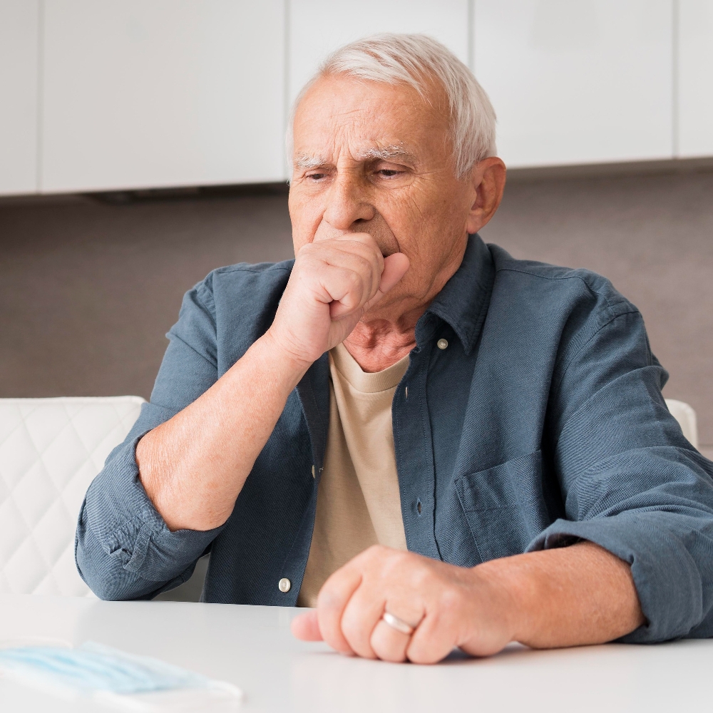 Older man with white hair coughing into his fist in a bright kitchen, a common early sign of stroke dysphagia 